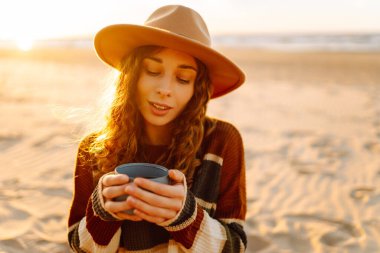 Genç bir kadın plajda piknikte oturup termostan sıcak bir içecek içiyor. Deniz manzarasının tadını çıkaran bir kız. Seyahat, hafta sonu, rahatlama ve yaşam tarzı konsepti