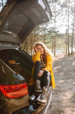 Genç bir kadın bagajda oturmuş doğanın tadını çıkarıyor. Arabayla yolculuk. Yaşam tarzı, seyahat, turizm, doğa konsepti.