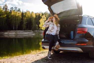 Genç bir kadın bagajda oturmuş doğanın tadını çıkarıyor. Arabayla yolculuk. Yaşam tarzı, seyahat, turizm, doğa konsepti.