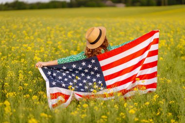 Tarlada rüzgarda Amerikan bayrağı tutan güzel bir genç kadın. Mavi gökyüzüne karşı yaz manzarası. Bağımsızlık Günü.