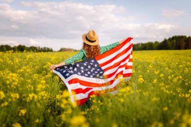 Tarlada rüzgarda Amerikan bayrağı tutan güzel bir genç kadın. Mavi gökyüzüne karşı yaz manzarası. Bağımsızlık Günü.