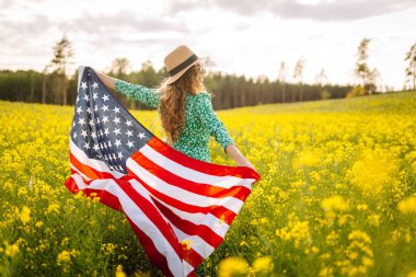 Tarlada rüzgarda Amerikan bayrağı tutan güzel bir genç kadın. Mavi gökyüzüne karşı yaz manzarası. Bağımsızlık Günü.
