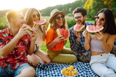 Parkta otururken meyve yiyen neşeli arkadaşlar. Gençler yaz tatilinde eğleniyor. Gıda, seyahat, doğa ve piknik konsepti