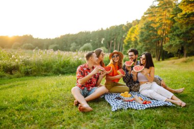 Parkta otururken meyve yiyen neşeli arkadaşlar. Gençler yaz tatilinde eğleniyor. Gıda, seyahat, doğa ve piknik konsepti
