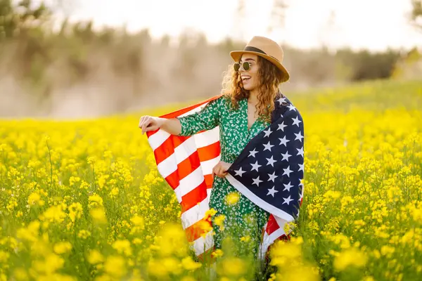 Tarlada rüzgarda Amerikan bayrağı tutan güzel bir genç kadın. Mavi gökyüzüne karşı yaz manzarası. Bağımsızlık Günü.