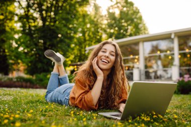 Laptop ve kablosuz kulaklık kullanırken parkta yeşil çimlerin üzerinde oturan çekici genç bir kadının portresi. Serbest Çalışan, Eğitim, İnternet, Teknoloji Konsepti.
