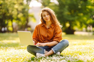 Laptop ve kablosuz kulaklık kullanırken parkta yeşil çimlerin üzerinde oturan çekici genç bir kadının portresi. Serbest Çalışan, Eğitim, İnternet, Teknoloji Konsepti.