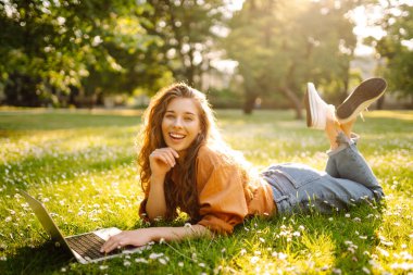 Laptop ve kablosuz kulaklık kullanırken parkta yeşil çimlerin üzerinde oturan çekici genç bir kadının portresi. Serbest Çalışan, Eğitim, İnternet, Teknoloji Konsepti.