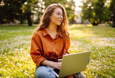 Laptop ve kablosuz kulaklık kullanırken parkta yeşil çimlerin üzerinde oturan çekici genç bir kadının portresi. Serbest Çalışan, Eğitim, İnternet, Teknoloji Konsepti.