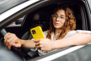 Sürücü koltuğunda oturan genç bir kadın akıllı telefona bakıyor, şehirde park ve gezinti için para ödüyor. Boş zaman, seyahat, teknoloji..