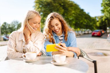 İki genç kadın kahve içiyor ve yazlık kafede otururken cep telefonu kullanıyor. Moda, güzellik, blog, turizm.