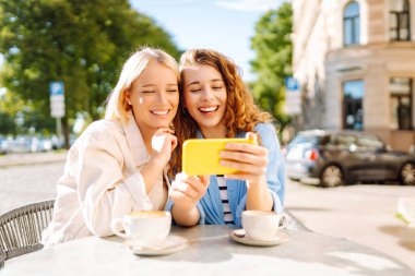 İki genç kadın kahve içiyor ve yazlık kafede otururken cep telefonu kullanıyor. Moda, güzellik, blog, turizm.