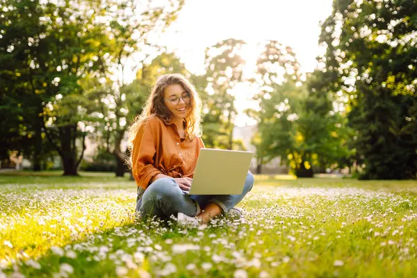 Laptop ve kablosuz kulaklık kullanırken parkta yeşil çimlerin üzerinde oturan çekici genç bir kadının portresi. Serbest Çalışan, Eğitim, İnternet, Teknoloji Konsepti.