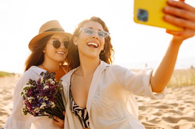Selfie zamanı. Gün batımında deniz kenarında poz veren iki genç kadın. Kızlar arkadaşına fotoğraf gösteriyor. Seyahat, blog, hafta sonu, rahatlama ve yaşam tarzı konsepti.