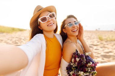 Selfie zamanı. Gün batımında deniz kenarında poz veren iki genç kadın. Kızlar arkadaşına fotoğraf gösteriyor. Seyahat, blog, hafta sonu, rahatlama ve yaşam tarzı konsepti.