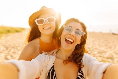Selfie zamanı. Gün batımında deniz kenarında poz veren iki genç kadın. Kızlar arkadaşına fotoğraf gösteriyor. Seyahat, blog, hafta sonu, rahatlama ve yaşam tarzı konsepti.