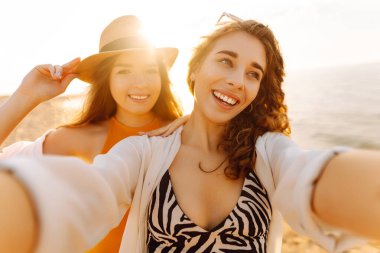 Selfie zamanı. Gün batımında deniz kenarında poz veren iki genç kadın. Kızlar arkadaşına fotoğraf gösteriyor. Seyahat, blog, hafta sonu, rahatlama ve yaşam tarzı konsepti.