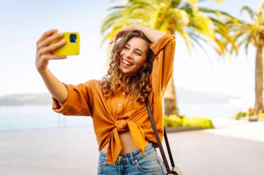 Genç bir kadın sokakta yürüyor ve akıllı telefon kamerasıyla selfie çekiyor. Güzel genç fotoğrafçı seyahat bloğu için fotoğraf çekiyor..