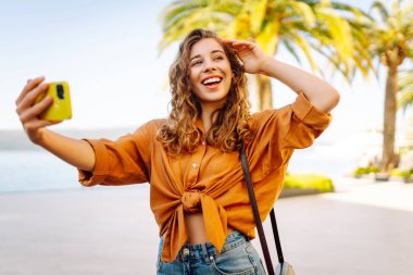 Genç bir kadın sokakta yürüyor ve akıllı telefon kamerasıyla selfie çekiyor. Güzel genç fotoğrafçı seyahat bloğu için fotoğraf çekiyor..