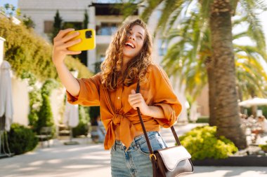 Genç bir kadın sokakta yürüyor ve akıllı telefon kamerasıyla selfie çekiyor. Güzel genç fotoğrafçı seyahat bloğu için fotoğraf çekiyor..