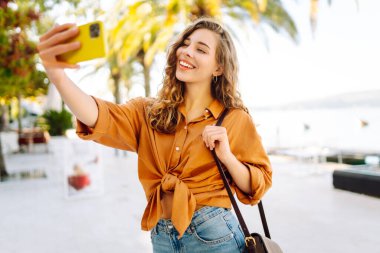 Genç bir kadın sokakta yürüyor ve akıllı telefon kamerasıyla selfie çekiyor. Güzel genç fotoğrafçı seyahat bloğu için fotoğraf çekiyor..