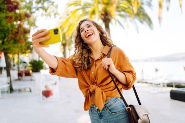 Genç bir kadın sokakta yürüyor ve akıllı telefon kamerasıyla selfie çekiyor. Güzel genç fotoğrafçı seyahat bloğu için fotoğraf çekiyor..