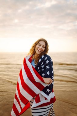 Ulusal Amerikan bayrağı taşıyan genç bir kadın yaz günbatımında okyanus sahillerinde tatil seyahatini kapsıyor. Amerika 'nın bağımsızlık günü. 4 Temmuz.