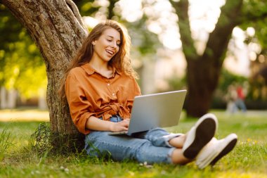 Dizüstü bilgisayarı olan genç bir kadın güneşli bir günde parkta çimlerin üzerinde oturur. Çevrimiçi eğitim, uzaktan çalışma. Eğitim çevrimiçi.