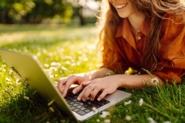 Dizüstü bilgisayarı olan genç bir kadın güneşli bir günde parkta çimlerin üzerinde oturur. Çevrimiçi eğitim, uzaktan çalışma. Eğitim çevrimiçi.