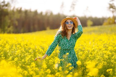 Şapkalı mutlu bir kadın çiçek açan bir kolza tarlasında yürüyor. Doğa, dinlenme. Yaz manzarası.