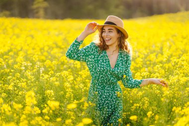 Şapkalı mutlu bir kadın çiçek açan bir kolza tarlasında yürüyor. Doğa, dinlenme. Yaz manzarası.