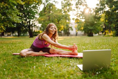 Genç bir kadının parkta dizüstü bilgisayarla yoga eğitimi var. Antrenman egzersizi. Sağlıklı yaşam tarzı, spor. Çevrimiçi eğitim.. 