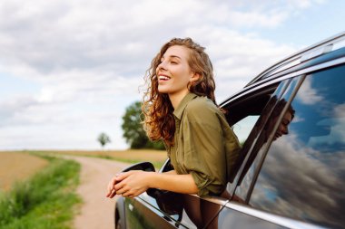 Elleri havada arabadan sarkan genç bir kadın doğada taze yaz rüzgarının tadını çıkarıyor. Yaz gezisi. Yaşam tarzı, seyahat, turizm, doğa, aktif yaşam