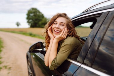 Elleri havada arabadan sarkan genç bir kadın doğada taze yaz rüzgarının tadını çıkarıyor. Yaz gezisi. Yaşam tarzı, seyahat, turizm, doğa, aktif yaşam