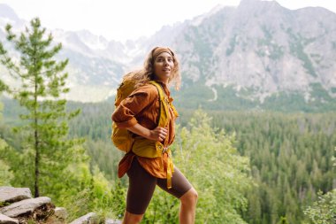 Sarı bir sırt çantasıyla gülümseyen dişi bir gezgin görkemli dağların güzel manzarasının tadını çıkarıyor. Seyahat, macera. Etkin bir yaşam biçimi kavramı.