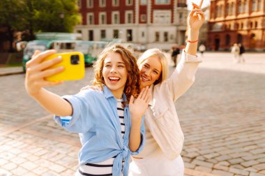 Yazlık kıyafetlerle gülümseyen iki genç kadın akıllı telefondan kendi portresini çekiyorlar. Sokakta poz veren mankenler. Moda, seyahat, teknoloji, blog konsepti.
