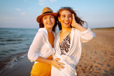 Gün batımında deniz kıyısında yürüyen iki genç bayan arkadaşın portresi. Seyahat, hafta sonu, rahatlama ve yaşam tarzı konsepti.