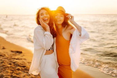 Gün batımında deniz kıyısında yürüyen iki genç bayan arkadaşın portresi. Seyahat, hafta sonu, rahatlama ve yaşam tarzı konsepti.