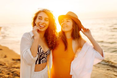 Gün batımında deniz kıyısında yürüyen iki genç bayan arkadaşın portresi. Seyahat, hafta sonu, rahatlama ve yaşam tarzı konsepti.