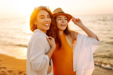 Gün batımında deniz kıyısında yürüyen iki genç bayan arkadaşın portresi. Seyahat, hafta sonu, rahatlama ve yaşam tarzı konsepti.