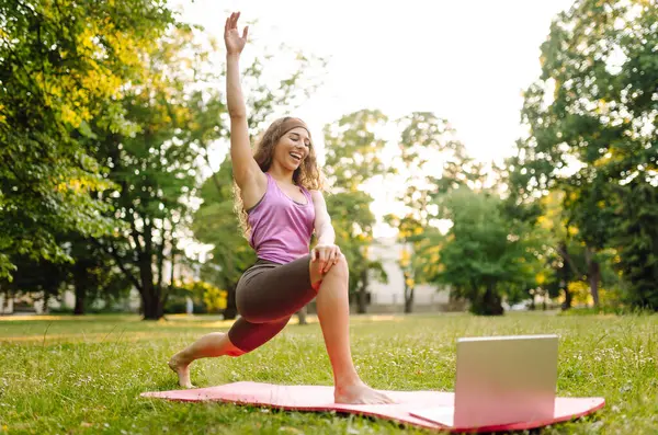 Genç bir kadının parkta dizüstü bilgisayarla yoga eğitimi var. Antrenman egzersizi. Sağlıklı yaşam tarzı, spor. Çevrimiçi eğitim.. 