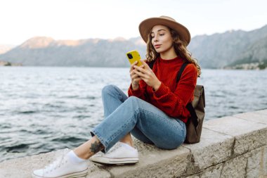 Sırt çantalı ve sıcacık giysili genç bayan gezgin gölün kenarında duruyor ve telefon ekranına bakıyor. Seyahat, yaşam tarzı, macera, konsept.