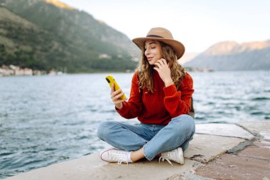 Sırt çantalı ve sıcacık giysili genç bayan gezgin gölün kenarında duruyor ve telefon ekranına bakıyor. Seyahat, yaşam tarzı, macera, konsept.