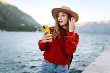 Sırt çantalı ve sıcacık giysili genç bayan gezgin gölün kenarında duruyor ve telefon ekranına bakıyor. Seyahat, yaşam tarzı, macera, konsept.