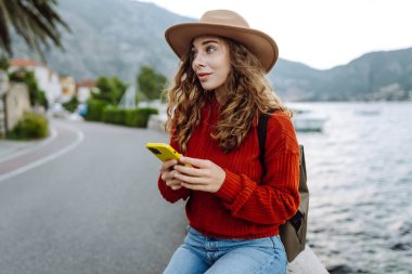 Sırt çantalı ve sıcacık giysili genç bayan gezgin gölün kenarında duruyor ve telefon ekranına bakıyor. Seyahat, yaşam tarzı, macera, konsept.
