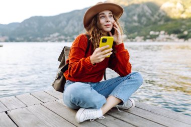 Sırt çantalı ve sıcacık giysili genç bayan gezgin gölün kenarında duruyor ve telefon ekranına bakıyor. Seyahat, yaşam tarzı, macera, konsept.