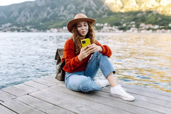 Sırt çantalı ve sıcacık giysili genç bayan gezgin gölün kenarında duruyor ve telefon ekranına bakıyor. Seyahat, yaşam tarzı, macera, konsept.