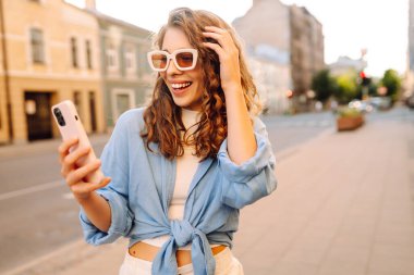 Sokakta elinde telefonla bekleyen güzel bir genç kadının fotoğrafı. Blogculuk, teknoloji, yaşam tarzı, seyahat, turizm, aktif yaşam