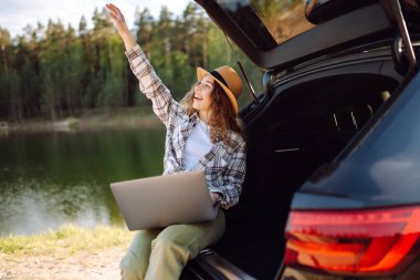 Dizüstü bilgisayar kullanan genç bir kadın doğada arabanın bagajında oturuyor. Seyahat ve teknoloji.