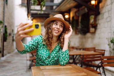 Genç bir kadın elinde dijital kamerayla yazlık kafede otururken selfie çekiyor. Moda, güzellik, blog, turizm.